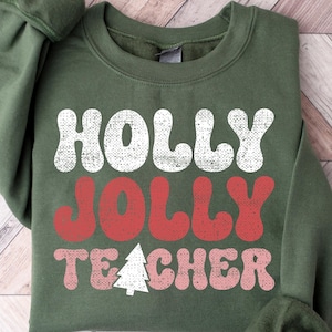 Op de afbeelding: Een groene sweatshirt met de tekst "HOLLY JOLLY TEACHER" in wit, rood en roze, met een witte kerstboomafbeelding. De sweatshirt wordt gepresenteerd op een houten oppervlak.