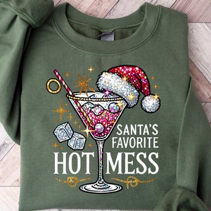 May include: Green sweatshirt with a festive design featuring a martini glass with a red drink, ice cubes, and a straw. The text "Santa's Favorite Hot Mess" is printed on the sweatshirt.