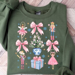 May include: A green sweatshirt with a Christmas-themed design featuring a Nutcracker, a ballerina, a Christmas tree, and pink bows.