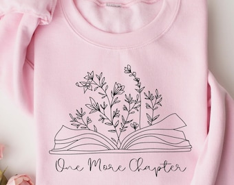 May include: A light pink sweatshirt with a black line drawing of an open book with flowers growing out of it. The text "One More Chapter" is below the book. The sweatshirt is folded on a white surface.