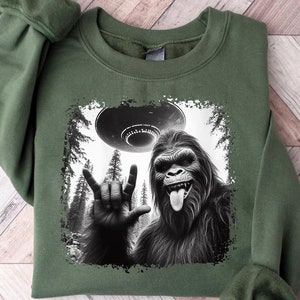 May include: Green sweatshirt with a black and white graphic print of a Bigfoot giving the rock and roll hand sign with a UFO in the background.