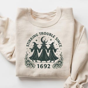 May include: Beige sweatshirt with a dark green graphic featuring three witches, a crescent moon, stars, and the text "Stirring Trouble Since 1692." The design is centered on the front of the folded garment.