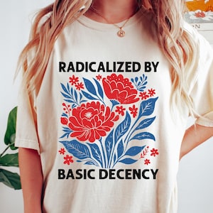 Op de afbeelding: Crèmekleurig T-shirt met een bloemenontwerp. Het ontwerp bevat rode bloemen, blauwe bladeren en de tekst "RADICALIZED BY BASIC DECENCY" in het zwart. Het T-shirt heeft een casual stijl.