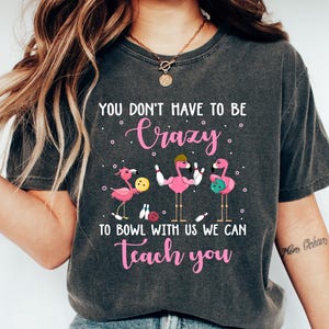May include: A gray t-shirt with the text "You don't have to be crazy to bowl with us we can teach you" in pink lettering. The design features three pink flamingos bowling.
