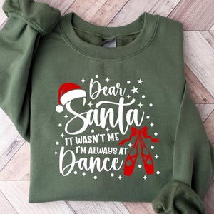 Könnte beinhalten: Ein olivgrünes Sweatshirt mit dem Text "Dear Santa, It wasn't me, I'm always at Dance" in Weiß, einem roten Weihnachtsmannhut und roten Ballettschuhen. Das Sweatshirt ist auf einer Holzoberfläche gefaltet.