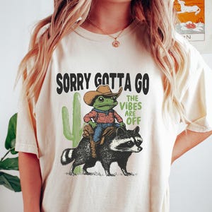 Può includere: T-shirt color crema con grafica di una rana con cappello da cowboy su un procione, con un cactus e la scritta "SORRY GOTTA GO THE VIBES ARE OFF".
