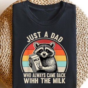 May include: Black t-shirt featuring a raccoon holding a milk carton, set against a retro sunset design. Text reads "JUST A DAD WHO ALWAYS CAME BACK WIHH THE MILK". The shirt is displayed on a woven, circular surface.