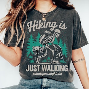 May include: Dark gray t-shirt with the text "Hiking is Just Walking Where You Might Die." The graphic features a skeleton hiker with a bear and pine trees. The shirt is a comfortable, casual style.