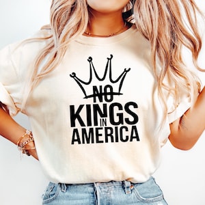 May include: Cream-colored t-shirt with the text "NO KINGS IN AMERICA" in bold black letters, topped with a crown graphic. The shirt is paired with light blue denim jeans.