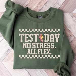 May include: An olive green sweatshirt with the text "TEST DAY NO STRESS. ALL FLEX." in a vintage font. A pink and yellow lightning bolt graphic is featured in the design. The sweatshirt is folded on a light wood grain surface.