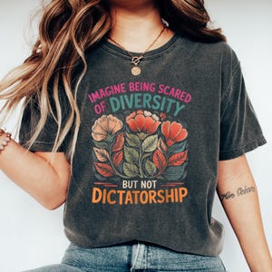 May include: Dark gray t-shirt with the text "IMAGINE BEING SCARED OF DIVERSITY BUT NOT DICTATORSHIP" above a floral design in orange, red, and pink. The shirt is paired with blue jeans and white sneakers.