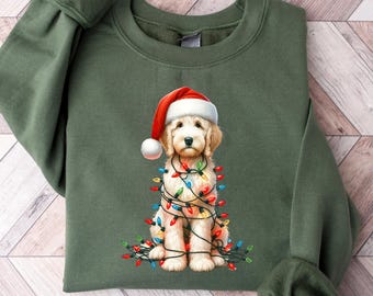 May include: An olive green sweatshirt featuring a graphic of a golden doodle wearing a Santa hat and wrapped in Christmas lights. The dog is light brown with a white face and a red and white Santa hat. The lights are multi-colored.