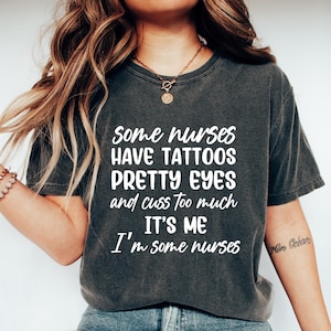 May include: Dark gray t-shirt with the text "Some nurses have tattoos, pretty eyes, and cuss too much. It's me, I'm some nurses" in white. The shirt is paired with blue jeans and white sneakers. A necklace and bracelet are also visible.