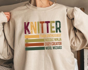 May include: A beige sweatshirt with the word "KNITTER" in colorful letters. Below the word are the phrases "Yarn Enthusiast", "Needle Ninja", "Cozy Creator", and "Wool Wizard" in a matching color scheme.