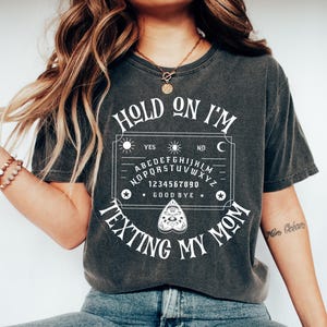 May include: A charcoal grey short-sleeve t-shirt featuring a white graphic of a spirit board with the text "HOLD ON I'M TEXTING MY MOM" printed in a stylized font. The design includes the alphabet, numbers zero through nine, and a planchette.