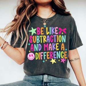 May include: A charcoal grey short-sleeve t-shirt featuring a colourful graphic print that reads BE LIKE SUBTRACTION AND MAKE A DIFFERENCE. The design includes bright pink, yellow, blue, and purple text, a pink smiley face, and star icons.