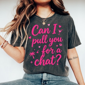 May include: A charcoal gray short-sleeve t-shirt featuring the bright pink text Can I pull you for a chat? along with graphic icons of a cocktail glass, a palm tree, hearts, stars, and lips. The shirt is paired with blue denim jeans and white sneakers.