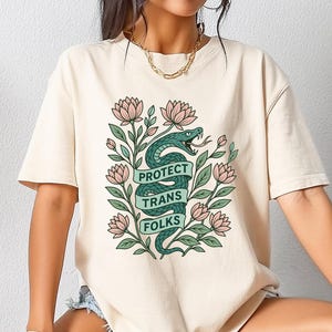 以下が含まれることがあります： クリーム色のTシャツ。緑色のヘビが「PROTECT TRANS FOLKS」と書かれたバナーを囲んでいます。デザインにはピンクの花と緑の葉が含まれています。このシャツはカジュアルな半袖スタイルです。