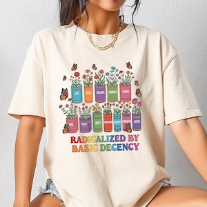 Könnte beinhalten: Cremefarbenes T-Shirt mit einem farbenfrohen Grafikdesign von Blumentöpfen mit Wörtern wie "Liebe" und "Gerechtigkeit". Der Satz "RADICALIZED BY BASIC DECENCY" ist darunter in Regenbogenfarben gedruckt. Schmetterlinge sind ebenfalls abgebildet.