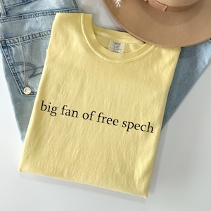 May include: A pale yellow t-shirt with the words "big fan of free spech" printed in black. The shirt rests on light blue denim jeans, with a gold necklace and a beige hat.