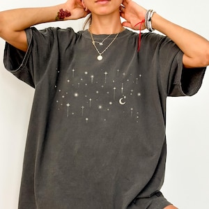 Könnte beinhalten: Dunkelgraues T-Shirt mit einem Sternen- und Mond-Design. Das übergroße T-Shirt hat kurze Ärmel und eine lockere Passform. Die Grafik ist in einem helleren Farbton gehalten, wodurch ein subtiler Kontrast entsteht. Ein lässiges und bequemes Oberteil.