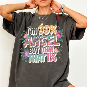 Op de afbeelding: Donkergrijs t-shirt met de tekst "I'm 99% ANGEL BUT OHH! THAT 1%" in kleurrijke, speelse letters. Het ontwerp bevat bloemaccenten in roze, paars en groen, wat zorgt voor een leuke en grillige uitstraling.