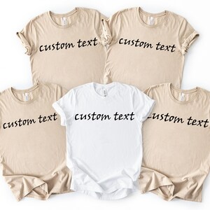 May include: Five t-shirts, four in a tan color and one in white, are displayed. Each shirt has the words "custom text" printed in a black, cursive font across the front. The shirts are laid out on a white surface.