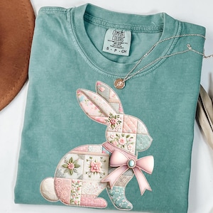 May include: A teal Comfort Colors t-shirt featuring a patchwork bunny design with a pink bow. The bunny is made of floral and patterned fabric squares. A silver necklace with a small pendant is draped across the shirt.