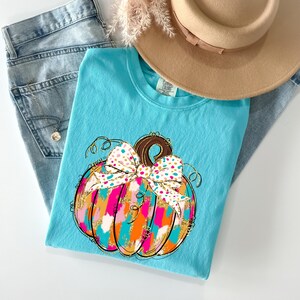 May include: A turquoise t-shirt featuring a colorful pumpkin design with a polka dot bow. The pumpkin has pink, orange, and teal brushstrokes with gold glitter accents. A tan hat and blue jeans are also in the image.