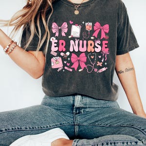May include: A dark gray t-shirt with a pink and white graphic design featuring the text "ER NURSE" and various medical symbols, including a stethoscope, bandages, and pills. The design also includes pink bows.