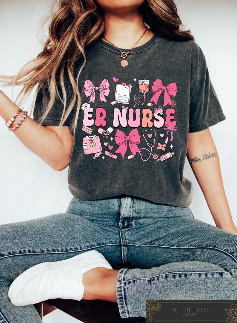 May include: A dark gray t-shirt with a pink and white graphic design featuring the text "ER NURSE" and various medical symbols, including a stethoscope, bandages, and pills. The design also includes pink bows.