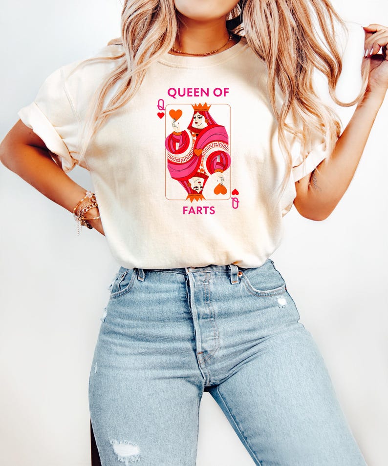 May include: Cream-colored t-shirt featuring a playing card design with the text "QUEEN OF FARTS." The card depicts a queen in pink and red with a crown and heart symbols. The shirt is paired with light blue jeans.