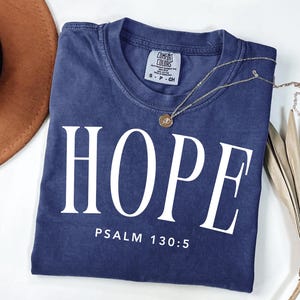 Puede incluir: Una camiseta azul marino con la palabra "HOPE" impresa en letras blancas grandes. Debajo de la palabra "HOPE" está el texto "PSALM 130:5". Un collar dorado con un colgante descansa sobre la camiseta. Un sombrero marrón y una planta seca están en el fondo.