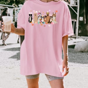 May include: Pink oversized t-shirt with a graphic of dogs wearing bunny ears and holding Easter eggs. The dogs are surrounded by flowers and other Easter decorations.