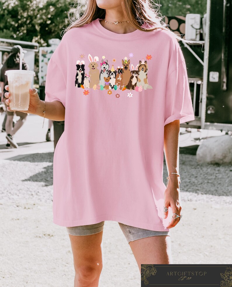May include: Pink oversized t-shirt with a graphic of dogs wearing bunny ears and holding Easter eggs. The dogs are surrounded by flowers and other Easter decorations.