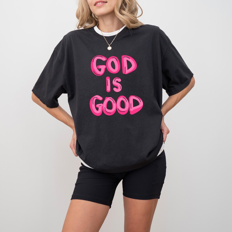 Pu&ograve; includere: T-shirt nero con la scritta "GOD IS GOOD" in lettere rosa a bolle. La maglietta ha un colletto bianco a girocollo e viene indossata con pantaloncini neri. &Egrave; visibile una collana d'oro.