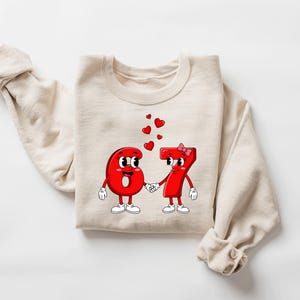 May include: A cream-colored sweatshirt featuring a graphic of the numbers 6 and 7 as cartoon characters holding hands, with red hearts floating above them. The numbers are red with white gloves and shoes. A pink bow adorns the number 7.