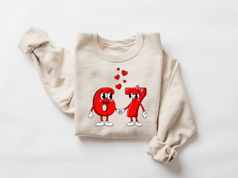 May include: A cream-colored sweatshirt featuring a graphic of the numbers 6 and 7 as cartoon characters holding hands, with red hearts floating above them. The numbers are red with white gloves and shoes. A pink bow adorns the number 7.
