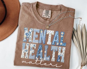 Könnte beinhalten: Ein braunes T-Shirt mit dem Schriftzug "MENTAL HEALTH matters" in einem Patchwork-Design aus blauen Blumen- und Vichy-Mustern. Eine silberne Halskette mit einem kleinen Anhänger liegt auf dem Shirt. Das Shirt ist auf einer weißen Oberfläche gefaltet.