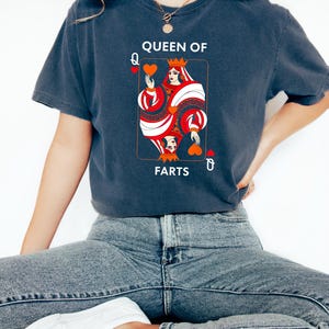May include: Dark blue t-shirt featuring a playing card design. The card depicts a queen holding hearts, with the text "QUEEN OF FARTS". The design is in red, white, and black. The shirt is a casual style.
