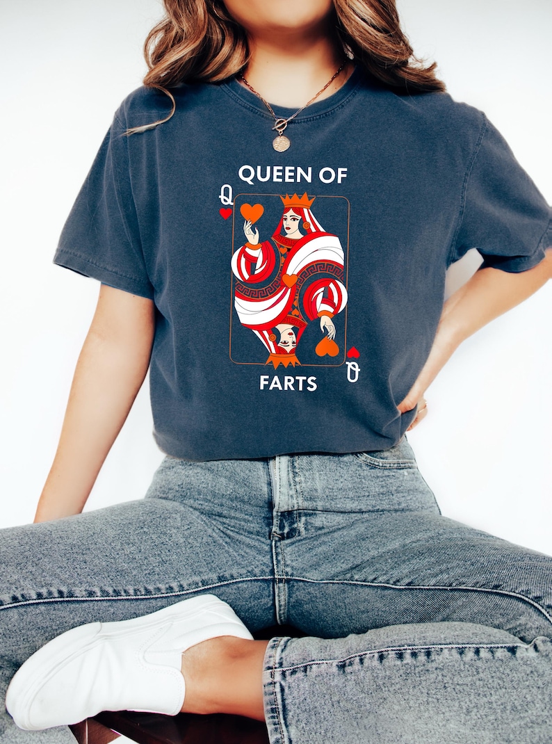 May include: Dark blue t-shirt featuring a playing card design. The card depicts a queen holding hearts, with the text "QUEEN OF FARTS". The design is in red, white, and black. The shirt is a casual style.