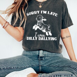 May include: Dark gray t-shirt with the text "SORRY I'M LATE I WAS DILLY DALLYING" and a white graphic of a leaping lamb. The shirt is a casual style, suitable for everyday wear.