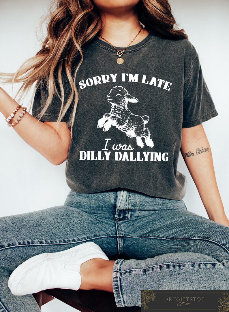 May include: Dark gray t-shirt with the text "SORRY I'M LATE I WAS DILLY DALLYING" and a white graphic of a leaping lamb. The shirt is a casual style, suitable for everyday wear.