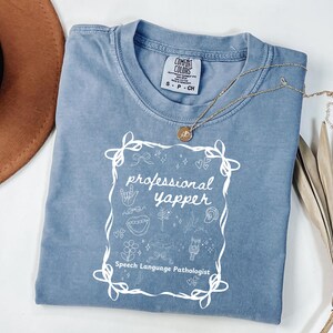 Puede incluir: Una camiseta azul Comfort Colors con las palabras "professional yapper" en blanco, junto con garabatos relacionados con la terapia del habla. Un collar dorado con un colgante descansa sobre la camisa. La camisa está doblada sobre una superficie blanca.