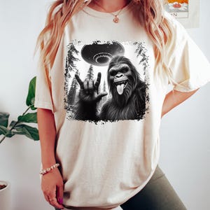 May include: A white t-shirt with a black and white graphic print of a Bigfoot giving the 'I love you' hand sign with a UFO in the background.