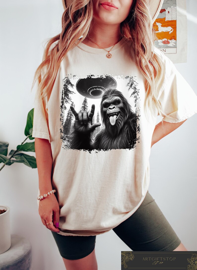 May include: A white t-shirt with a black and white graphic print of a Bigfoot giving the 'I love you' hand sign with a UFO in the background.