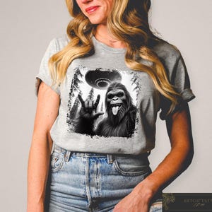 May include: A gray t-shirt with a black and white graphic print of a Bigfoot giving the peace sign with a UFO in the background.