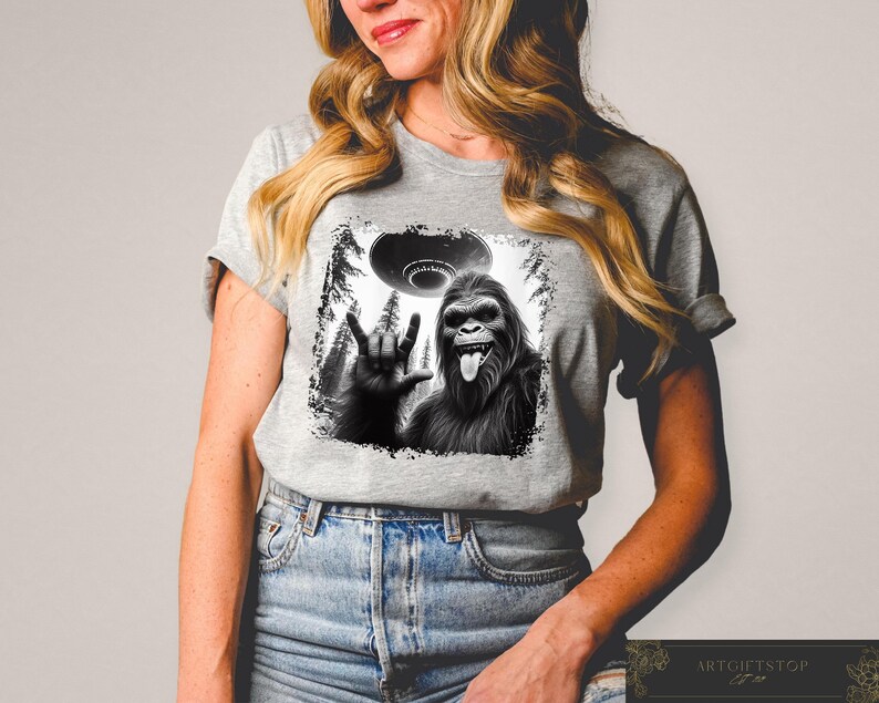May include: A gray t-shirt with a black and white graphic print of a Bigfoot giving the peace sign with a UFO in the background.