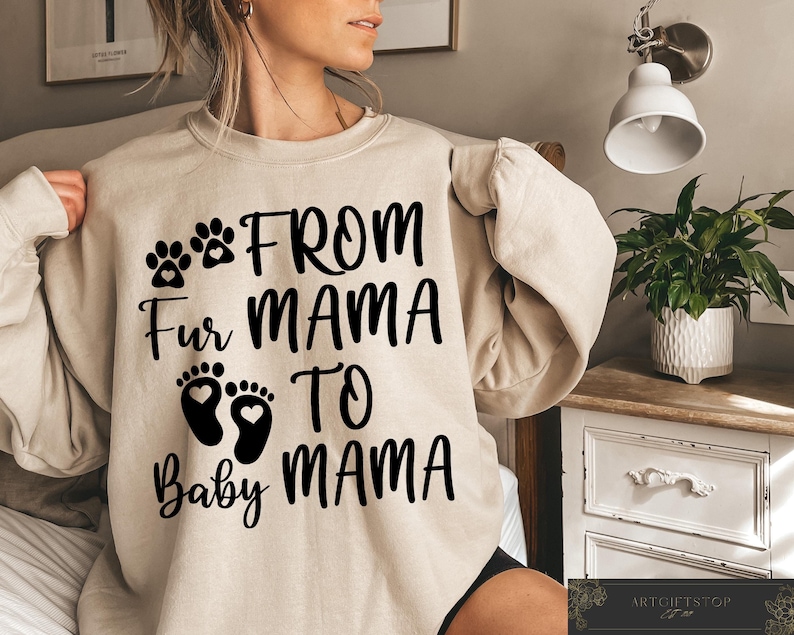 May include: Beige sweatshirt with the text "From Mama Fur Mama To Baby Mama" in black. The design includes paw prints, baby footprints with hearts, and text. The sweatshirt is a neutral color and has long sleeves.