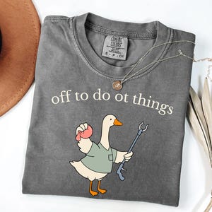 May include: A charcoal gray t-shirt featuring the white text "off to do ot things" above a cartoon goose wearing a sage green shirt. The goose holds a pink object in one wing and a blue reaching tool in the other. The shirt has a Comfort Colors label.